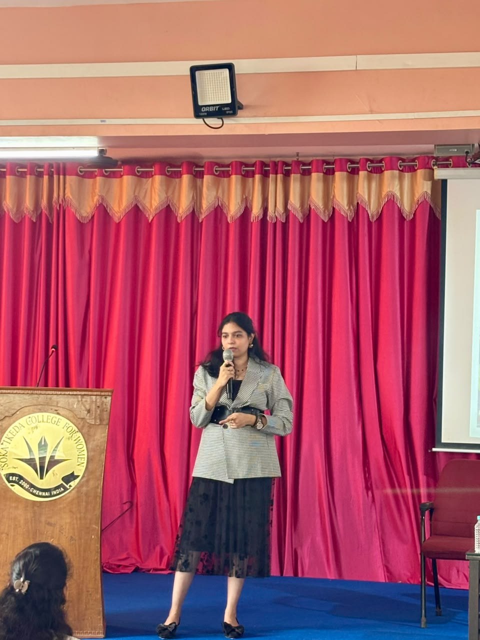 Naveena Srinivasan speaking on stage in front of students